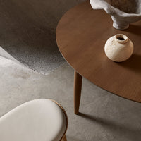 Round brown wooden table with minimalist design, paired with a white upholstered chair, set against a neutral backdrop.