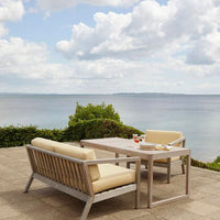 Cream-colored cushions on wooden outdoor furniture with a serene coastal backdrop and clear blue sky.
