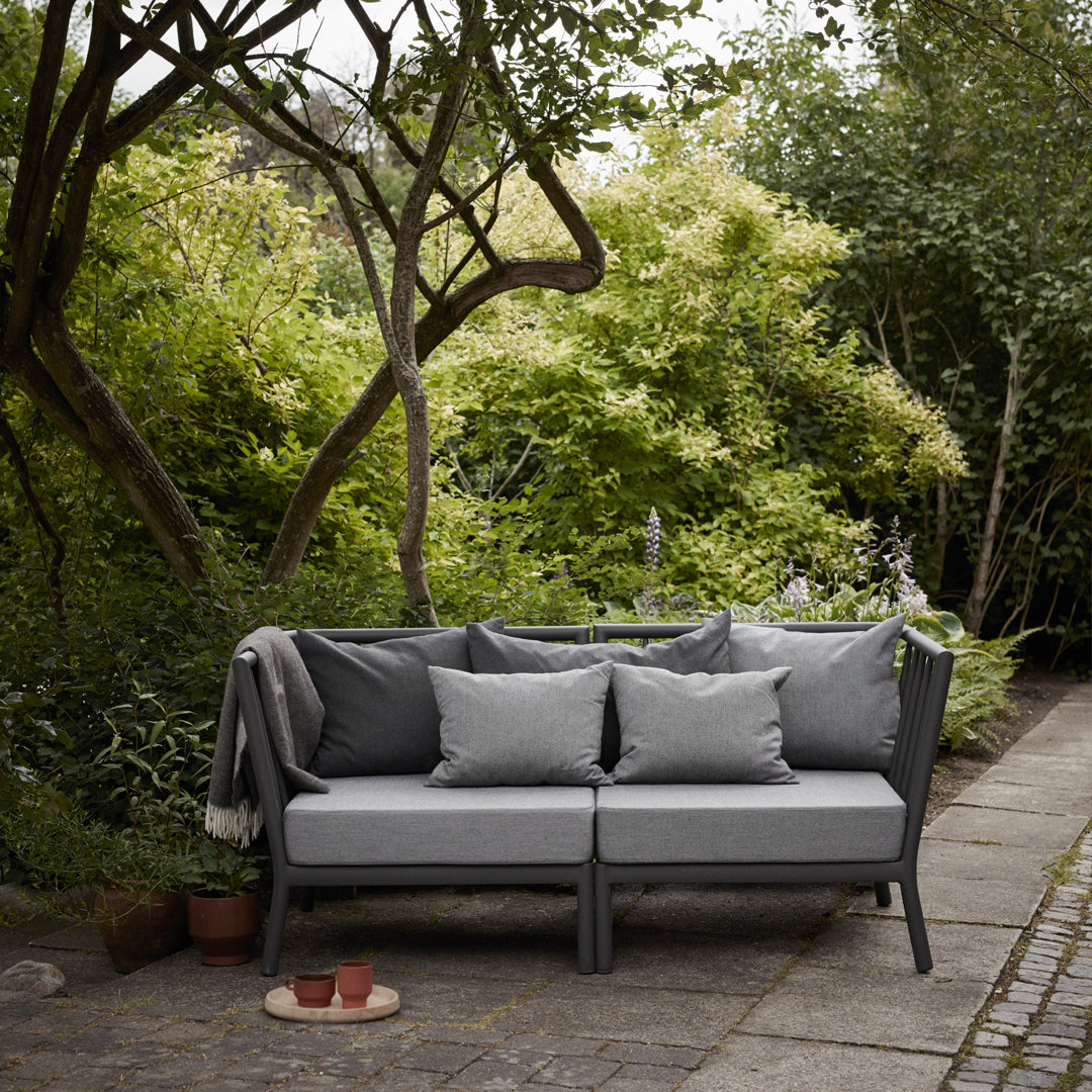 Gray outdoor sofa with matching pillows and a sleek design, set against a lush green garden backdrop.