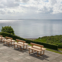 Light wood benches with natural grain patterns on a stone patio overlooking a calm blue sea and green foliage.