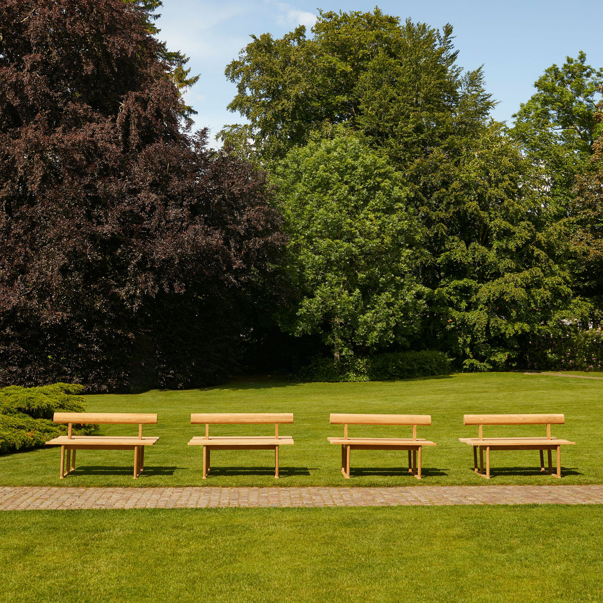"Five natural wood benches on a green lawn, surrounded by lush trees and blue skies."