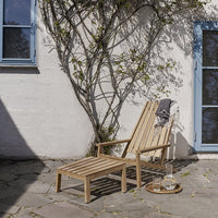 "Natural wood lounge chair with a slatted design, set against a textured stone and white wall backdrop."