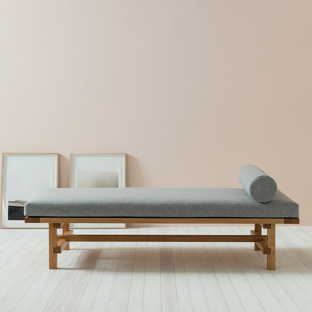 Gray fabric chaise lounge with a round bolster, set against a soft pink wall and light wood floor. Minimalist design.