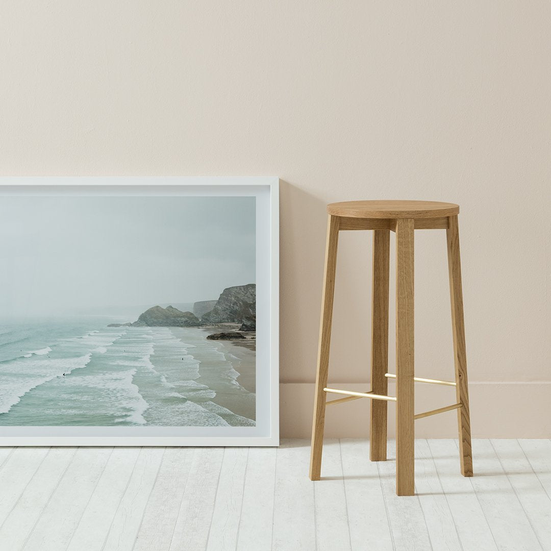 Natural wood bar stool against a soft beige wall with a coastal landscape print, featuring light blue and white ocean waves.