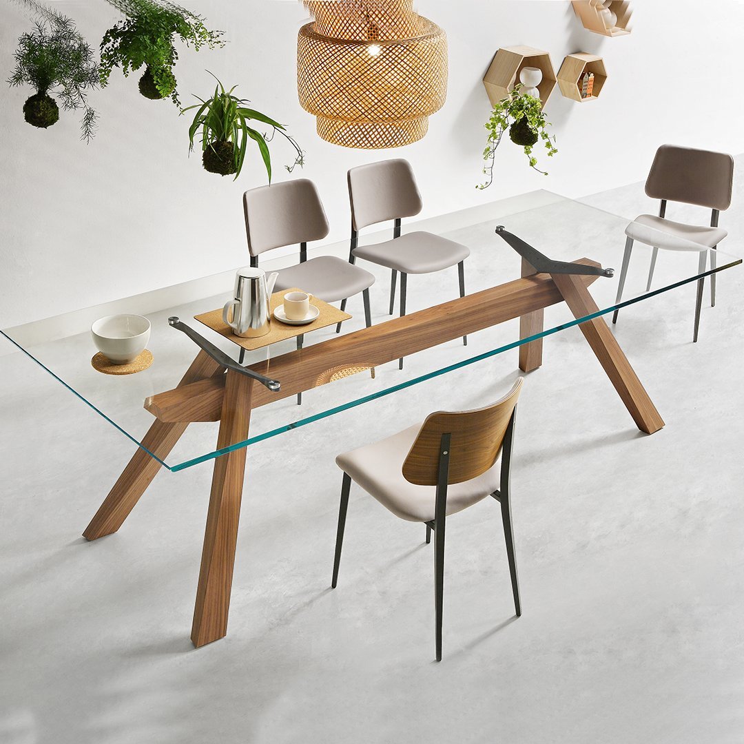 Elegant glass dining table with a light wooden base, surrounded by beige and gray upholstered chairs. Modern, minimalistic design.