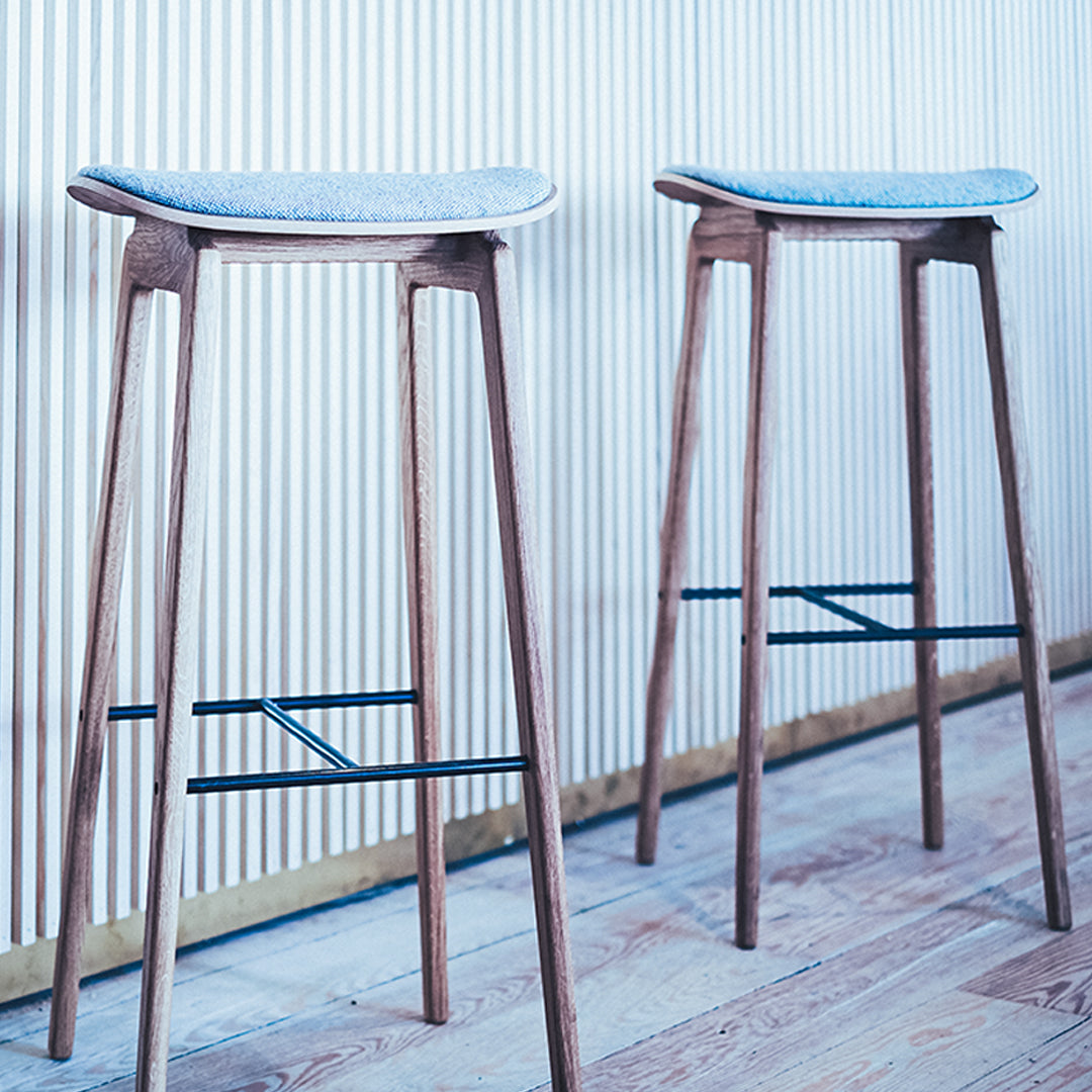Natural wood bar stools with light blue patterned cushions and sleek black footrests against a vertical ribbed wall.
