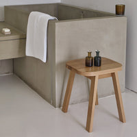 Natural wood stool with a light oak finish, paired with gray concrete and white towel accents.