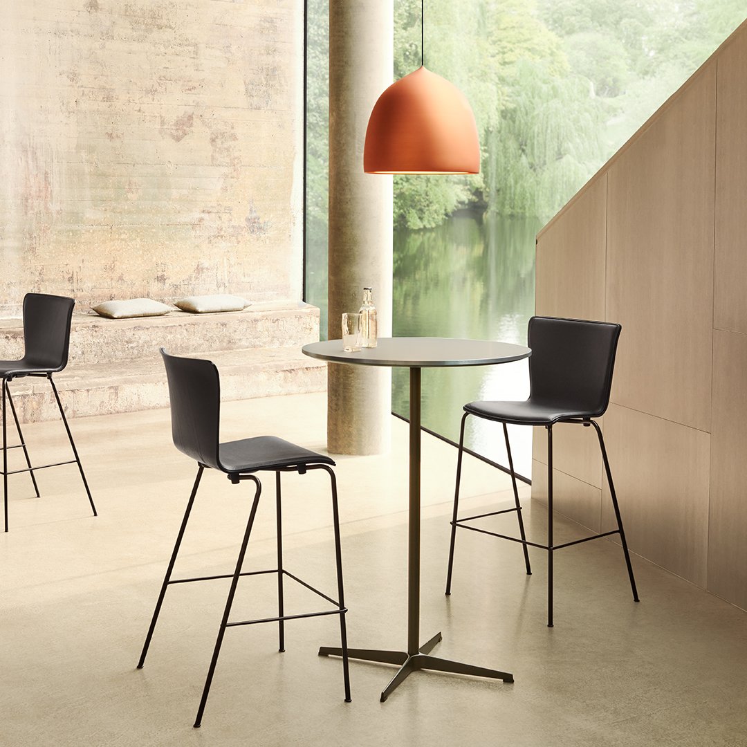 Minimalist black stool and round table with gold base, complemented by a soft orange pendant light. Neutral backdrop.