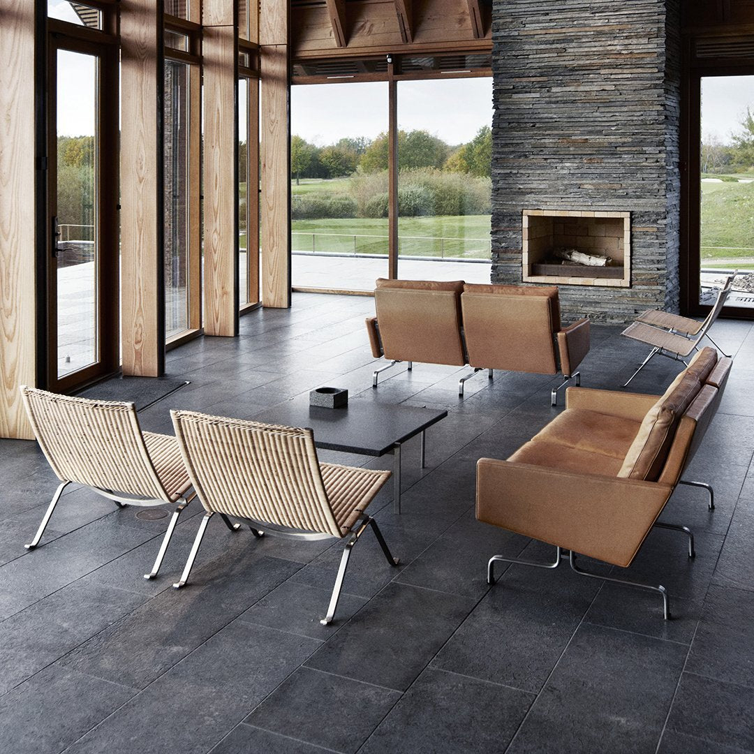 Modern living room with sleek brown leather furniture and woven chairs on dark slate flooring, surrounded by large windows.