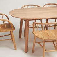 Natural wood dining table with a smooth oval top, paired with simple, light wooden chairs featuring classic slatted backs.