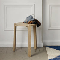 Light oak stool with a simple design, featuring a stack of textured gray, brown, and earthy-toned fabrics on top.