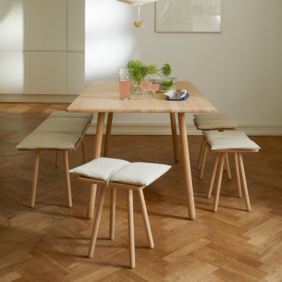 Natural wood dining table with light beige cushions on minimalist benches, set against warm oak flooring.