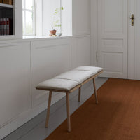 White padded bench with light wood legs on a textured brown rug, set against a neutral wall.