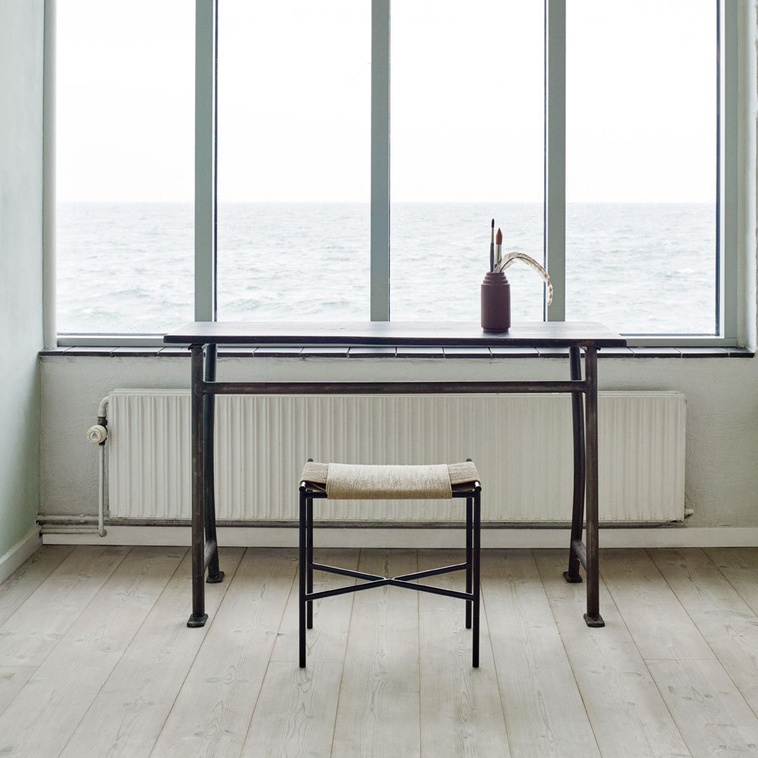Minimalist black metal desk with a beige patterned stool, set against light wood flooring and bright, airy windows.