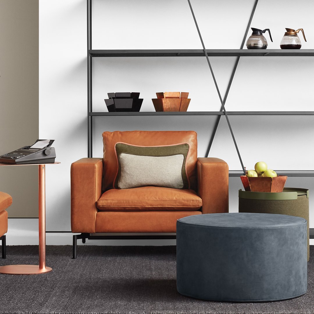 Rust orange leather chair with a green and cream pillow, gray ottoman, and earthy green accents on shelves.