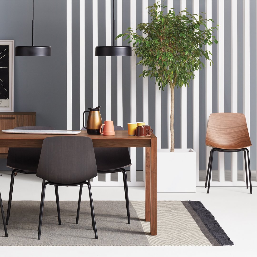 Wooden dining table with black and gray chairs, accented by vibrant orange and yellow mugs, set against striped gray and white walls.