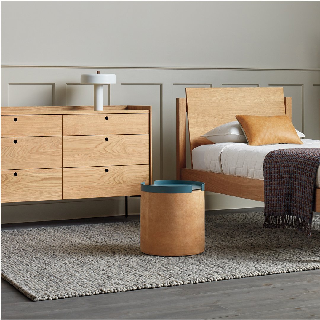Natural oak furniture with warm wood tones, soft beige bedding, and a textured gray rug accented by a blue and wood storage bin.