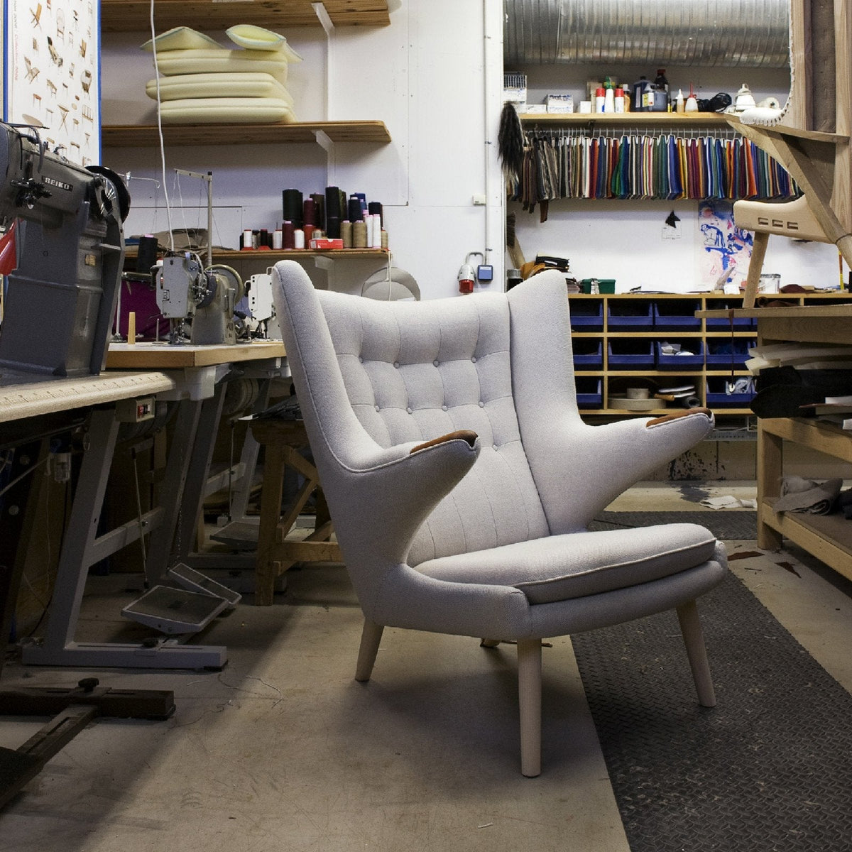 Light gray armchair with tufted upholstery, set in a workshop with wooden accents and colorful fabric swatches.