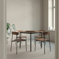 Modern round wooden table with black metal legs, complemented by four minimalist brown chairs, in a neutral-toned room.