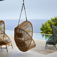 "Hanging rattan chair with a natural finish, showcasing an intricate woven pattern against a serene coastal backdrop."