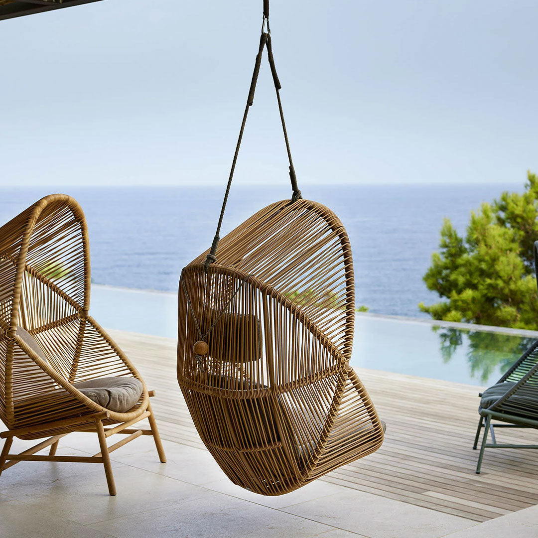 Hanging rattan chair in natural bamboo finish, featuring an elegant, woven pattern against a serene coastal backdrop.