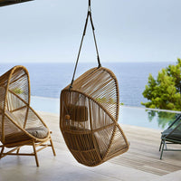 Hanging rattan chair in natural bamboo finish, featuring an elegant, woven pattern against a serene coastal backdrop.