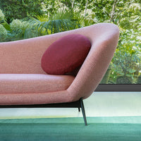 Pink upholstered sofa with a round burgundy pillow, set against a green carpet and lush greenery outside.