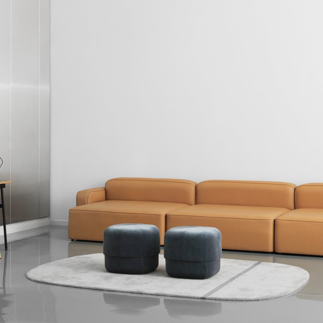 Tan sectional sofa with gray ottomans on a round white rug in a modern, minimalist space.