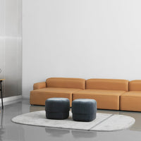Tan sectional sofa with gray ottomans on a round white rug in a modern, minimalist space.