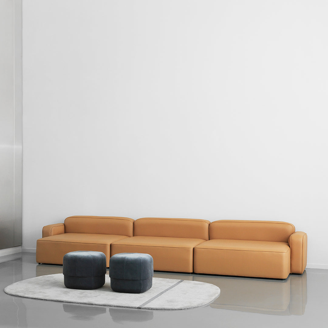Modern minimalistic sofa in warm caramel color with smooth lines, complemented by soft gray round ottomans and a textured rug.