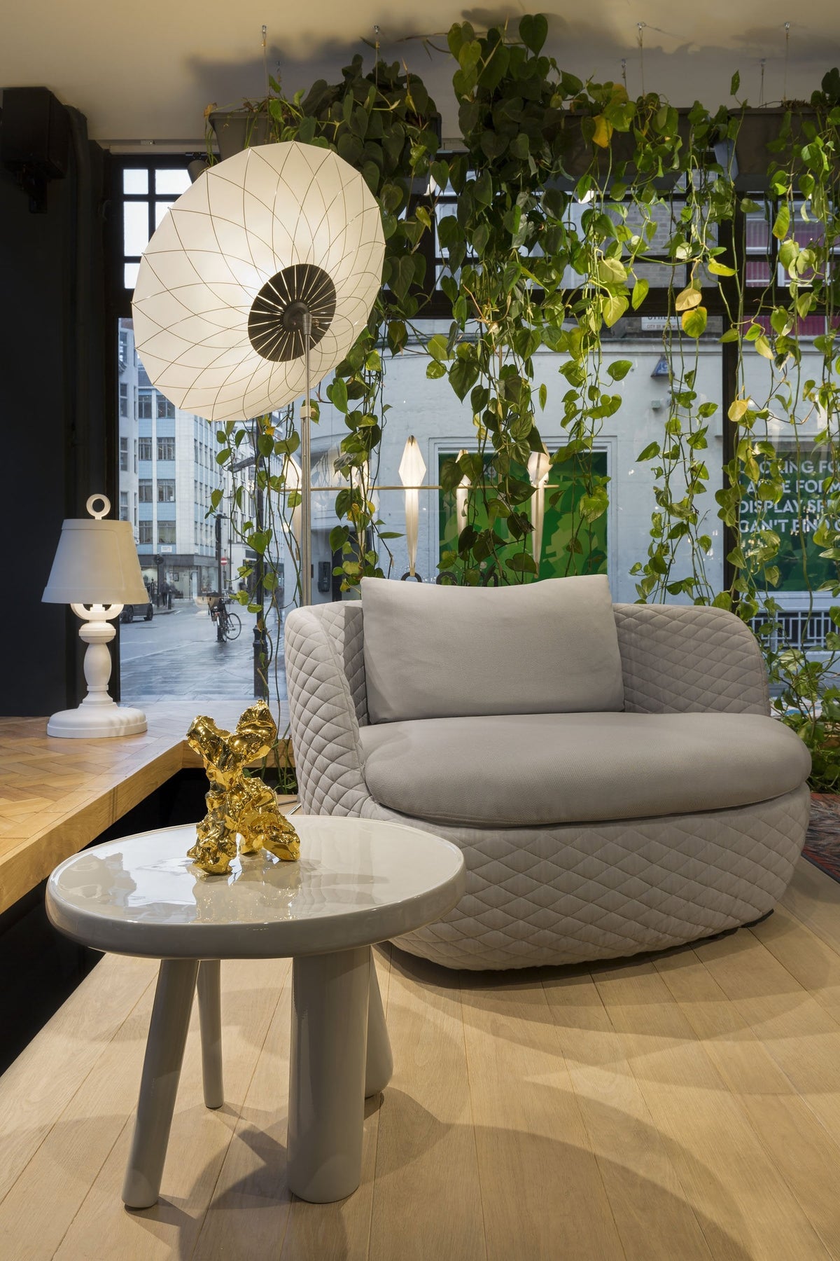Gray curved sofa with textured pattern, white lamp, and minimalist gray table, surrounded by green foliage.