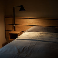 Wooden bed frame with soft gray bedding, illuminated by warm light from a stylish black lamp.