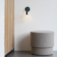 Green wall sconce with a clear bulb beside a light gray, textured upholstered stool in a minimalist setting.