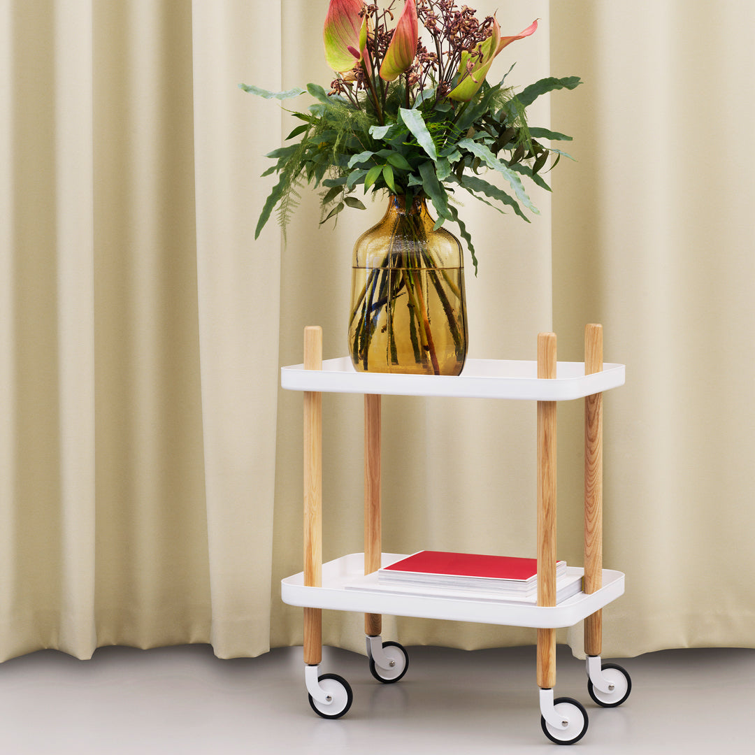 White two-tier cart with wooden legs and a pop of red, featuring a floral arrangement in a yellow vase.