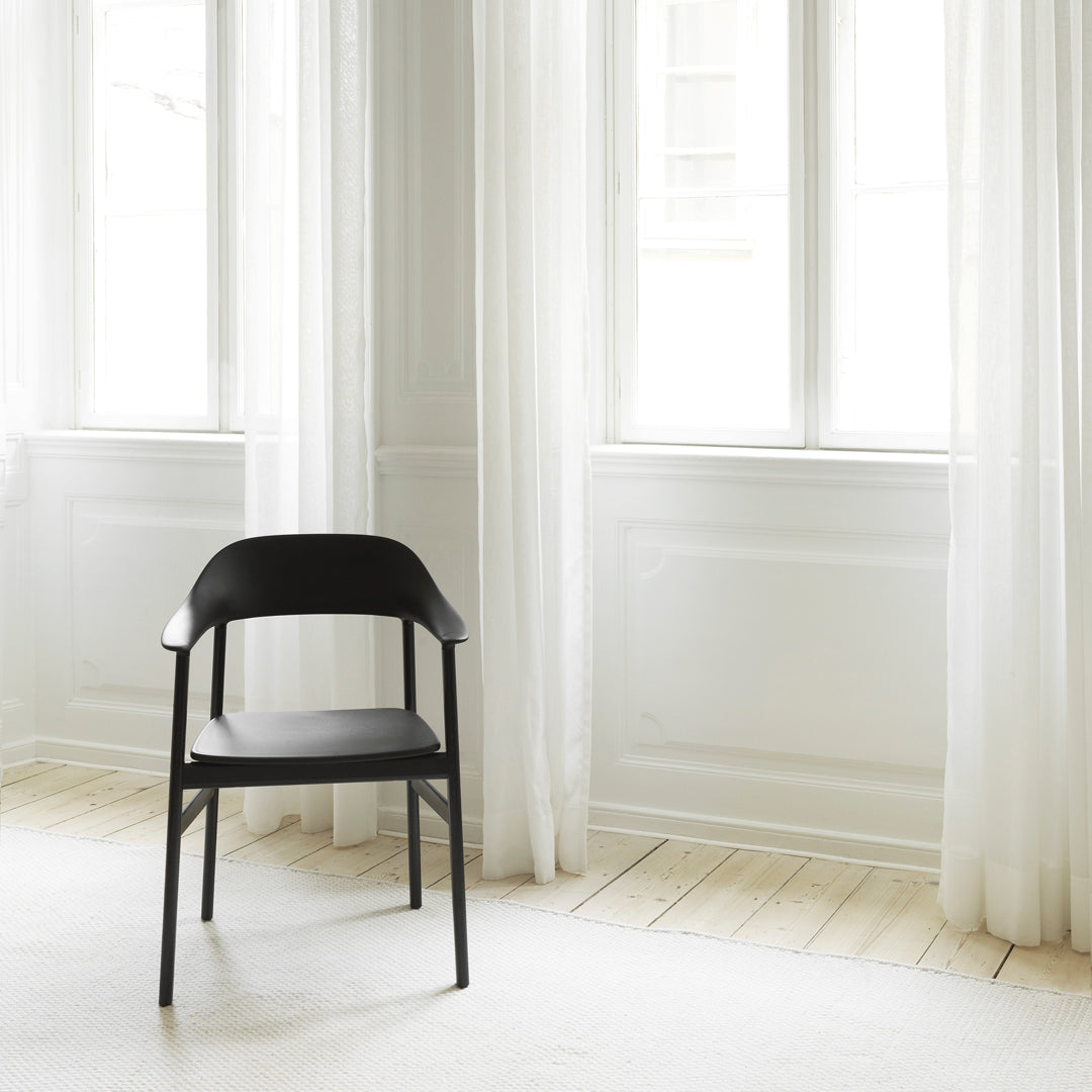 Black chair with a sleek design against a light, airy room with sheer white curtains and a neutral wooden floor.