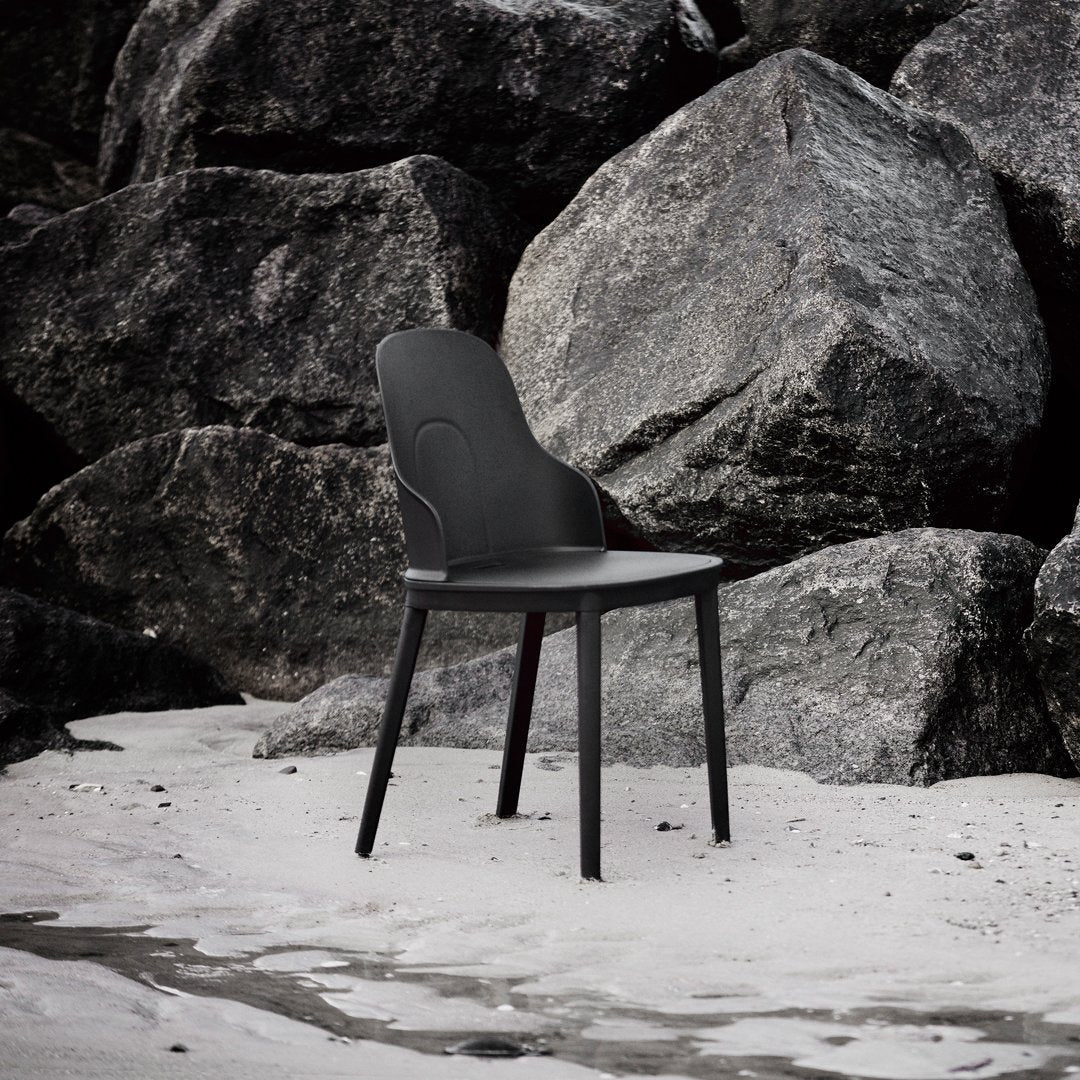 Black minimalist chair with smooth curves, set against rugged gray rocks and sandy beach, highlighting modern design.