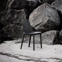 Black minimalist chair with smooth curves, set against rugged gray rocks and sandy beach, highlighting modern design.
