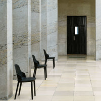 Three minimalist black chairs aligned on a light tiled floor, against textured stone walls and a dark door.