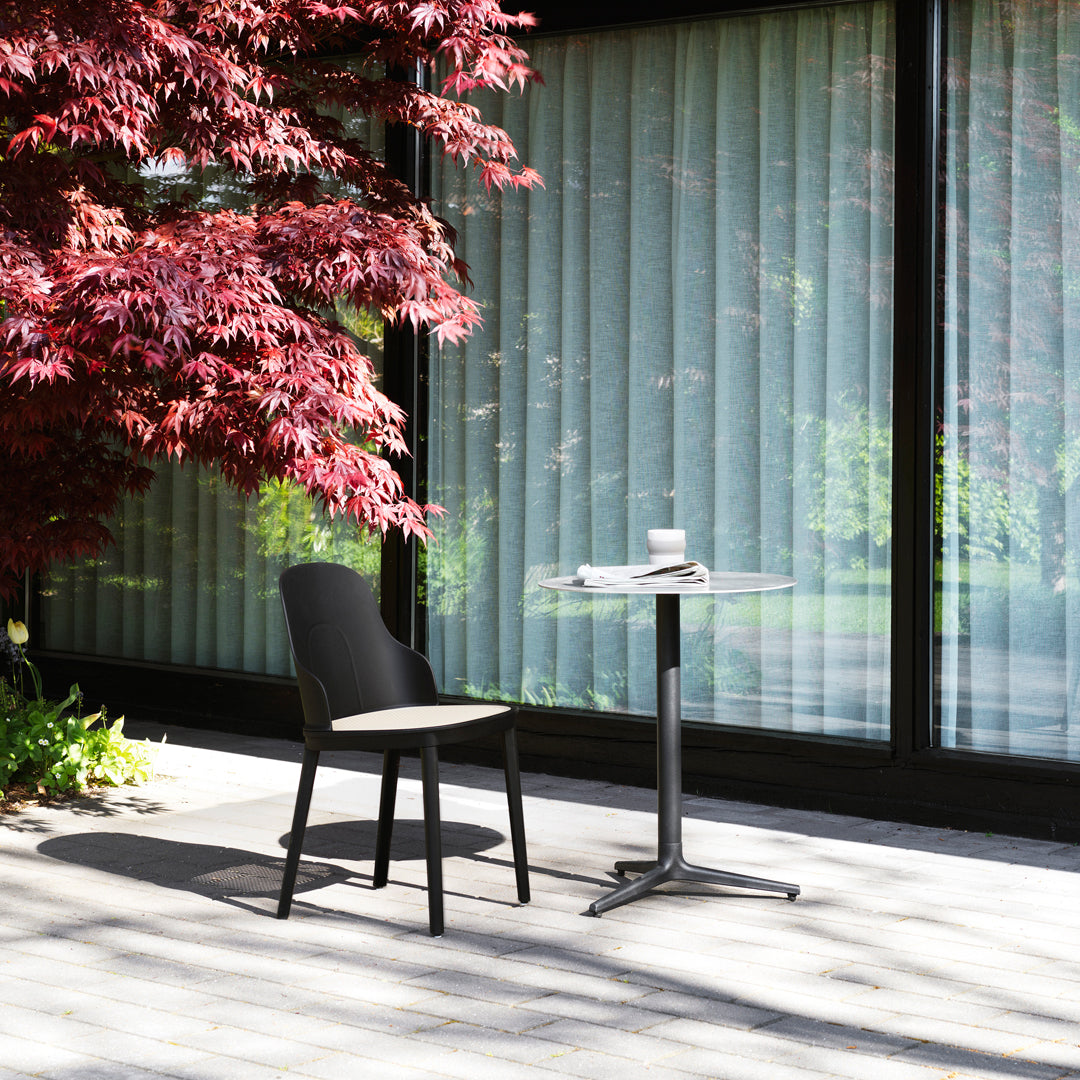 Black chair and sleek glass table on a patio, accented by a vibrant red maple tree and soft beige tiles.