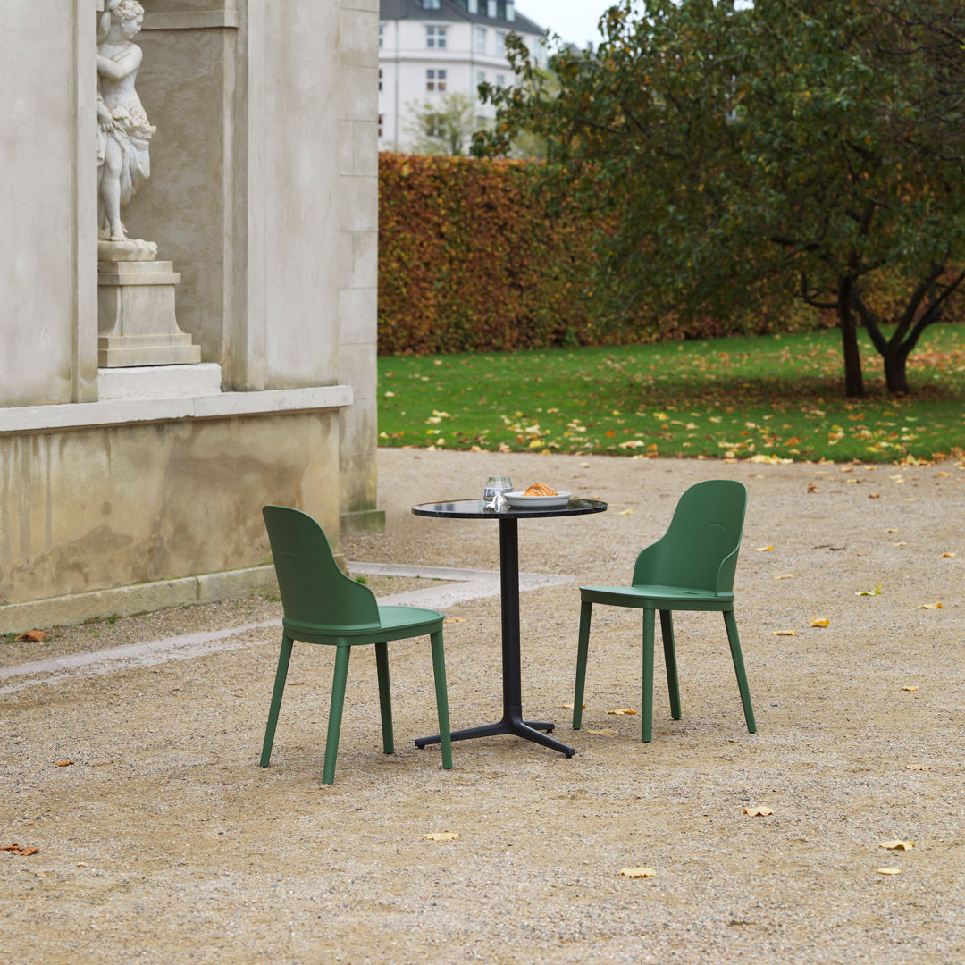 Green plastic chairs paired with a black table, set against a neutral stone backdrop and autumn foliage.
