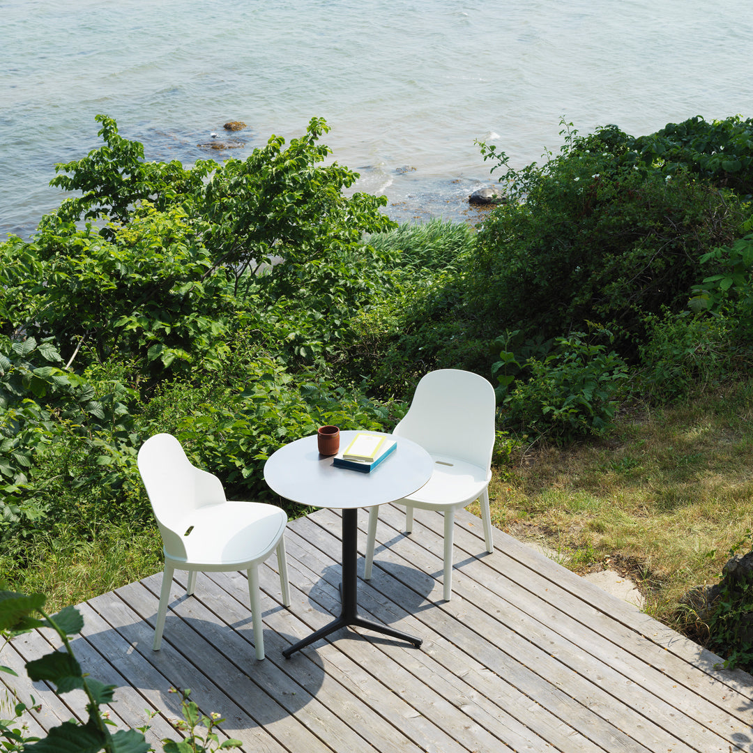 "White outdoor table and chairs against lush greenery by the water, featuring minimalist design with a black base."