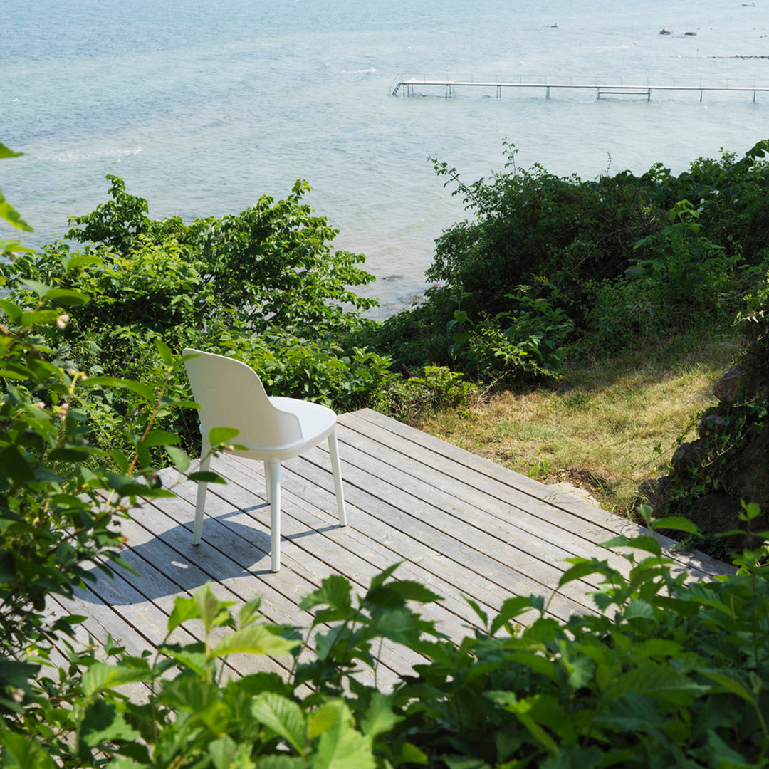 White chair on wooden deck, surrounded by lush green foliage and serene blue water in the background.