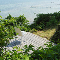 White chair on wooden deck, surrounded by lush green foliage and serene blue water in the background.