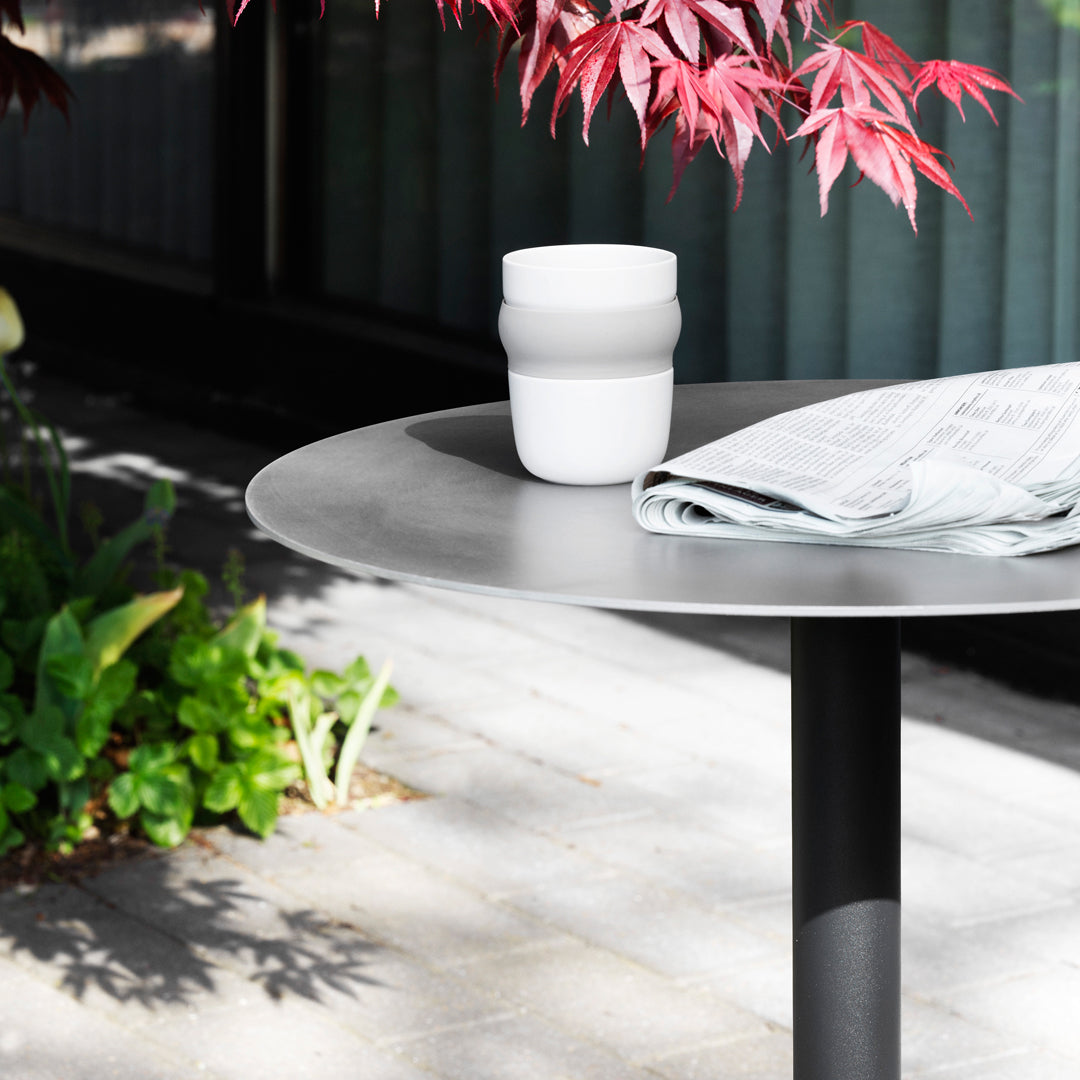 Stylish black round table with a white cup and soft grey napkin, surrounded by vibrant green plants and red leaves.