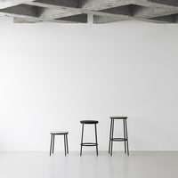 Three minimalist bar stools in black and natural wood against a clean white backdrop with a concrete ceiling.