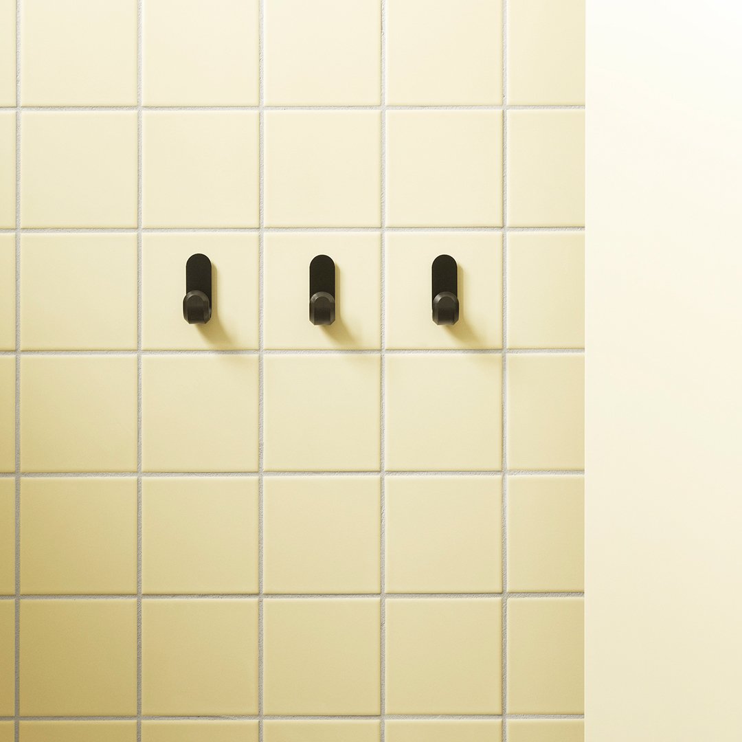 Black hooks on a pale yellow tiled wall, featuring a subtle grid pattern and smooth light yellow surface.
