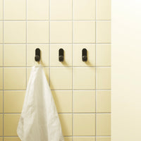 Black hooks on a light yellow tiled wall, with a white towel hanging from one hook. Simple and modern design.