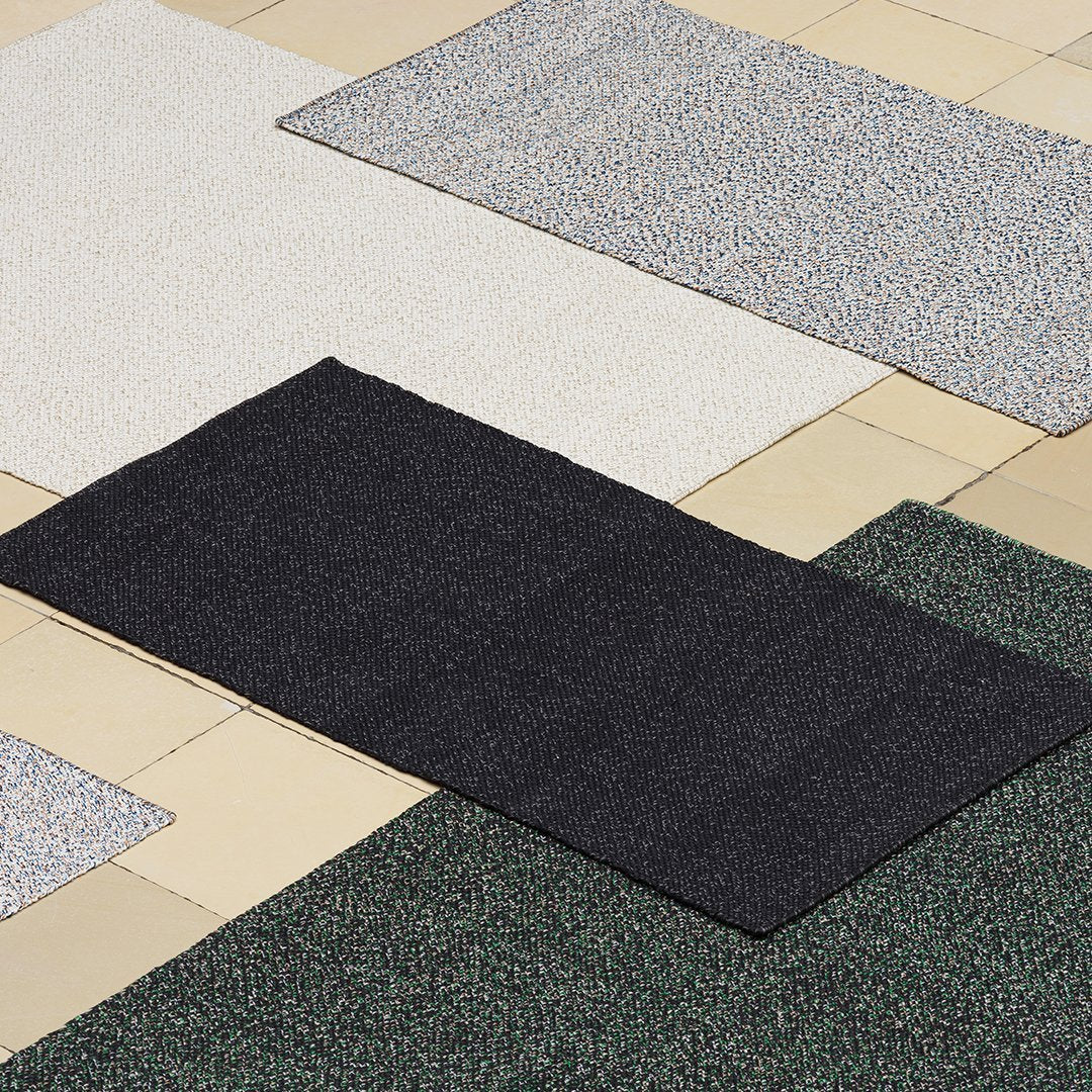 Various textured rugs in black, green, gray, and white colors, arranged on a light tiled surface.