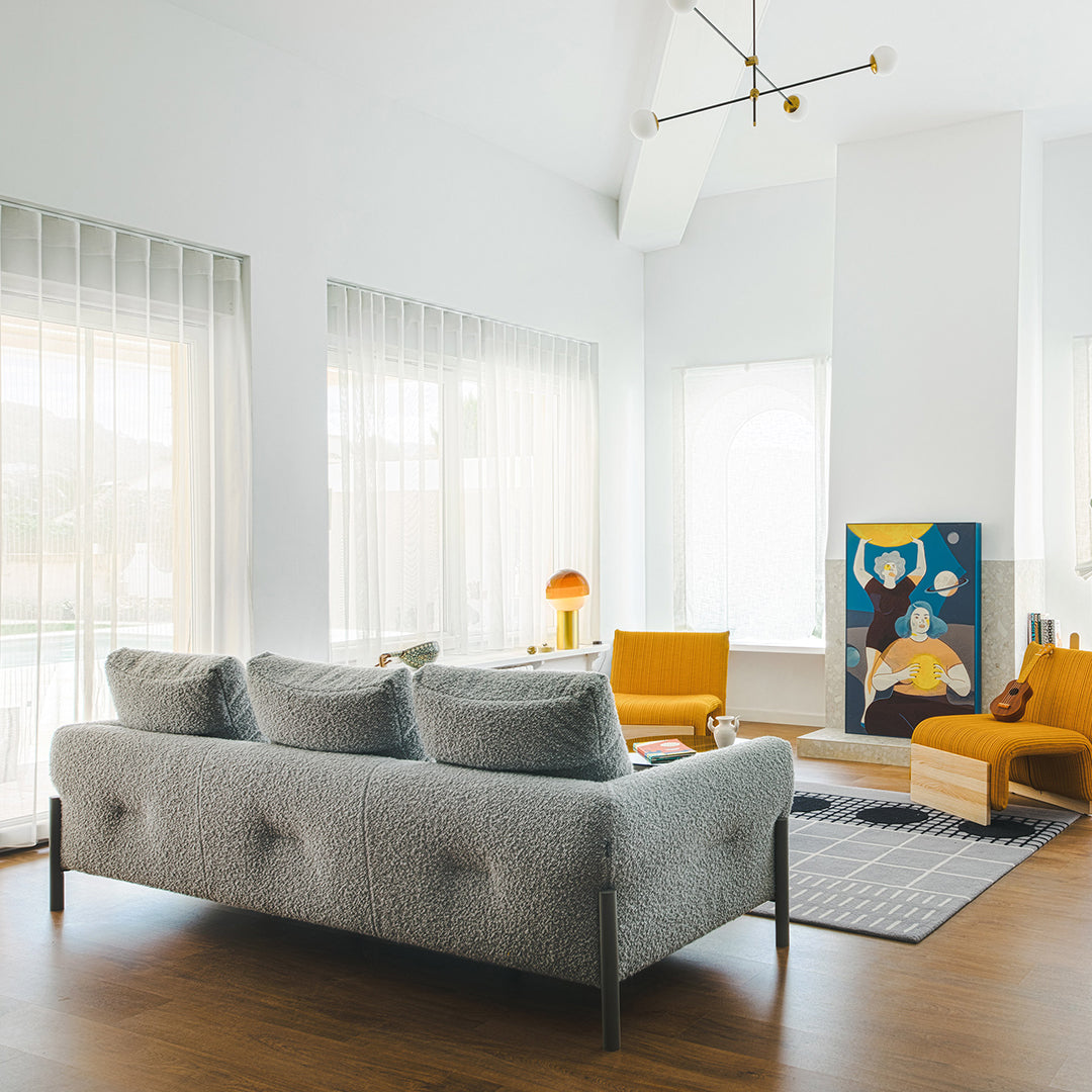 Gray tufted sofa, mustard yellow accent chairs, soft blue artwork, and geometric patterns on a light rug in a modern living room.