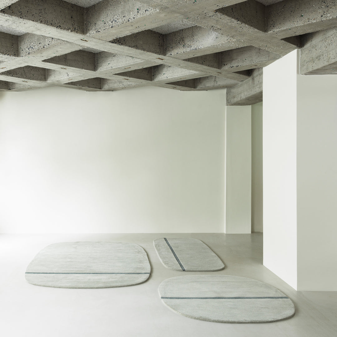 Abstract light gray rugs with soft curves and subtle blue lines, set against a minimalist, neutral backdrop.
